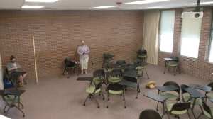 A woman sitting at a desk in the corner while another stands against the wall. Desks are spaced 6 feet apart along the walls. Other desks are scattered in the foreground.