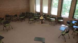 A woman in a sun hat crouching down and holding a long stick on the floor between two desks