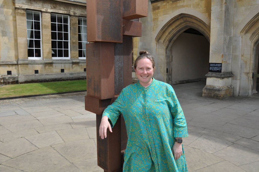 Photo of a white woman with dark hair in a high bun, wearing a high-necked, long-sleeved green dress. She is standing outdoors, leaning on a blocky brass sculpture, with stone pavement and buildings behind her.